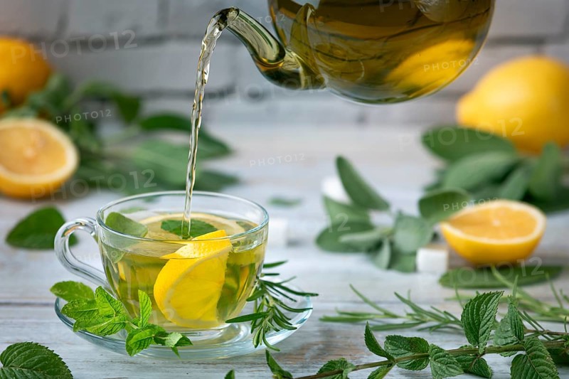 Tea is poured from a glass jug into a glass cup, surrounded by lemons and herbs