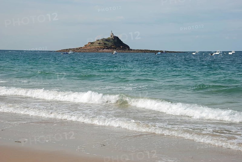 A lighthouse standing on an island surrounded by the ocean, with waves crashing on a sandy beach and boats floating nearby, Erquy, Les Hopitaux