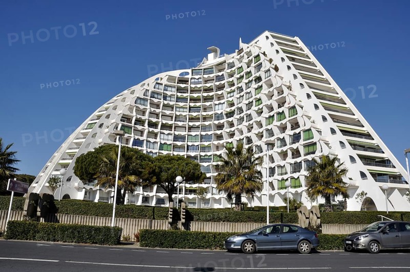A modern curved building with a white facade and numerous balconies, set in an urban environment with trees and parked cars under a clear sky, Buildings in La Grande Motte