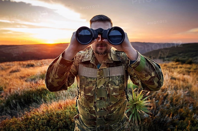 Man in camouflage clothing looks with binoculars over a green landscape at sunset, AI generated, AI generated