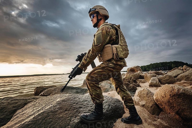 A soldier in uniform and helmet with a weapon stands on rocks by the ocean under a stormy sky, AI generated, AI generated