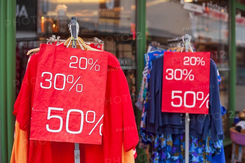 Red sign discount on a shopping bag in front of a clothing rack with t-shirts and shop window, Germany, Europe