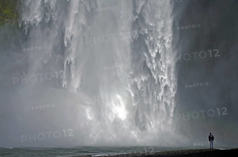 A man stands in front of an impressive waterfall, surrounded by splashing spray, Skogafoss, Iceland, Europe