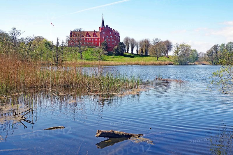 A red castle on a hill on the shore of a tranquil lake, surrounded by nature, Traeneker Slot, Manor Route, Langeland, Denmark, Europe