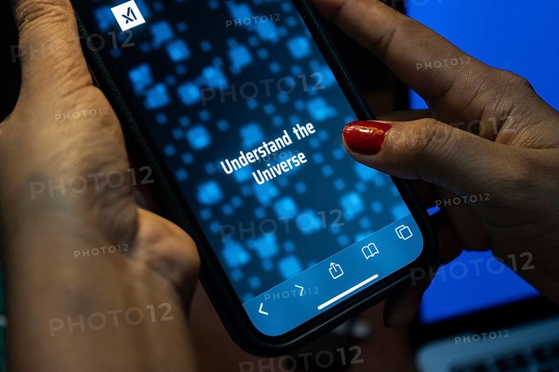 In this photo illustration, A person browses website of xAI, displayed on a mobile phone, Elon Musk images in the background screen, in Guwahati, India 0n 15 July 2023. Tesla CEO Elon Musk launches Artificial Intelligence company xAI on 12 July 2023, the company's goal is to understand the true nature of the universe