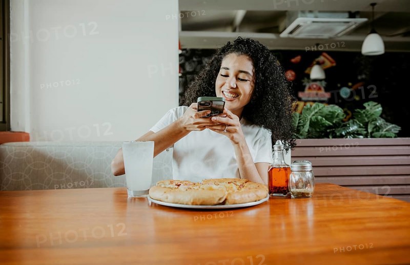 Afro girl taking a picture of a pizza with the phone in a pizzeria. Smiling young woman using phone photographing a pizza in a restaurant