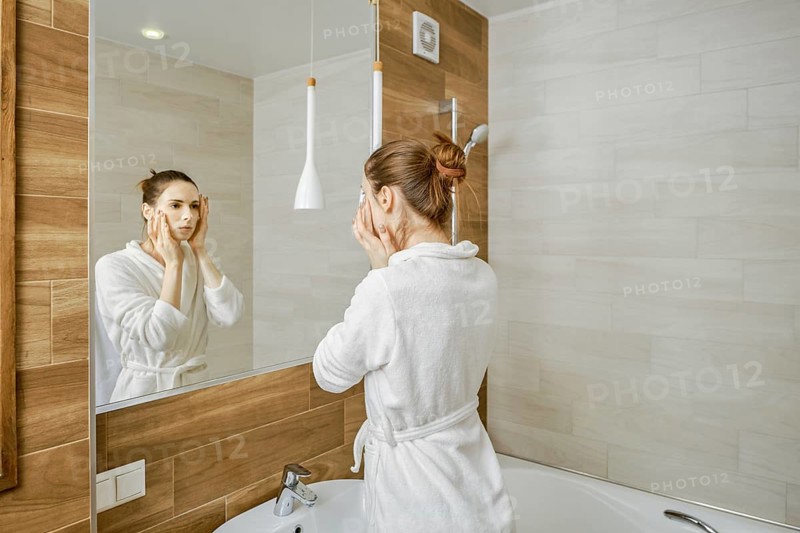 A woman in a white robe is washing her face, standing before a large mirror in a well-lit, contemporary bathroom