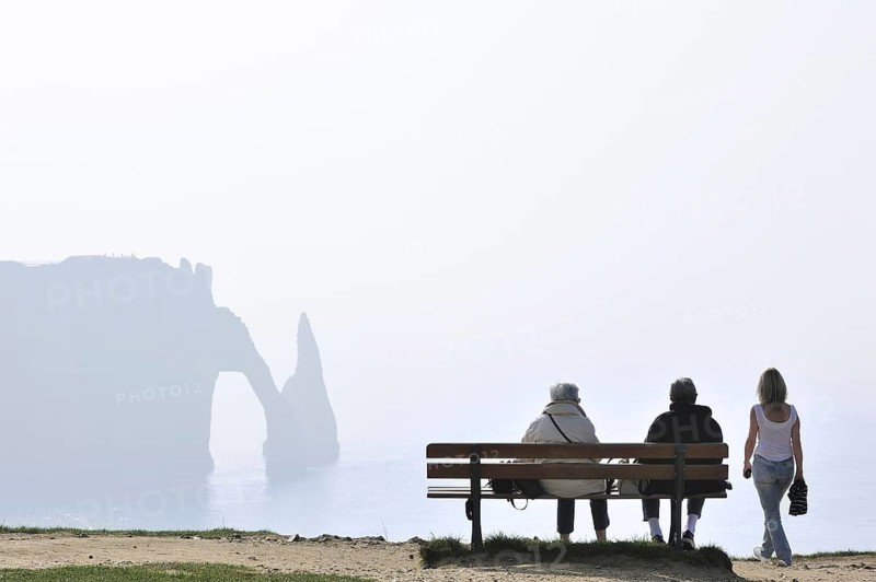 Elderly tourists sitting on bench looking at the Porte D'Aval, a natural arch in the chalk cliffs at Etretat, Côte d'Albâtre, Upper Normandy, France, Europe