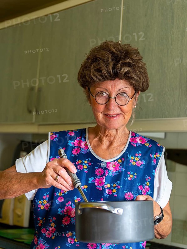 Granny with old smock apron, glasses and wig in the kitchen, holding cooking pot and ladle, Germany, Europe