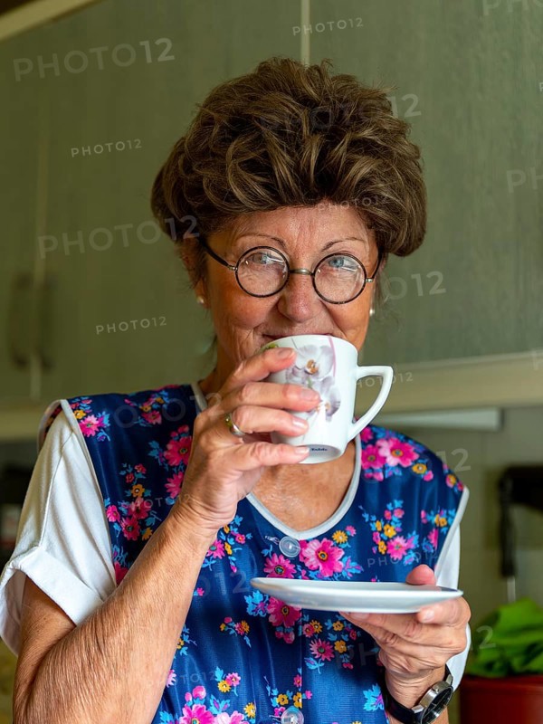 Granny with old smock apron, glasses and wig in the kitchen, holding coffee cup, Germany, Europe