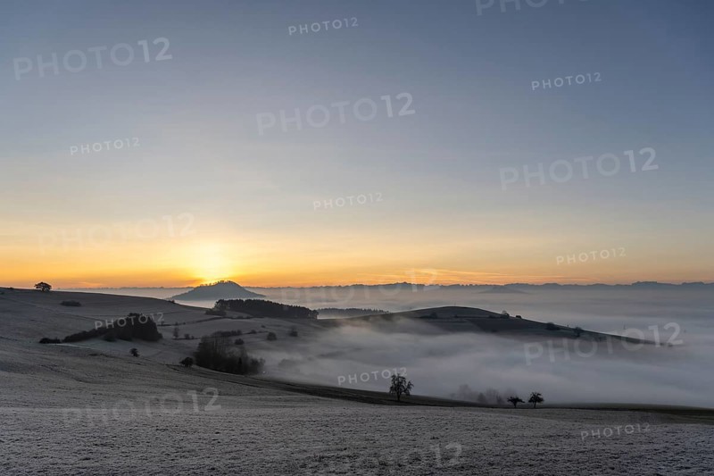 View from the old post road near Leipferdingen into the wintry and misty Hegaulandschaft shortly in front of sunrise on the horizon of the Hohenhewen and the Alpine chain, district of Constance, Baden-Württemberg, Germany, Europe