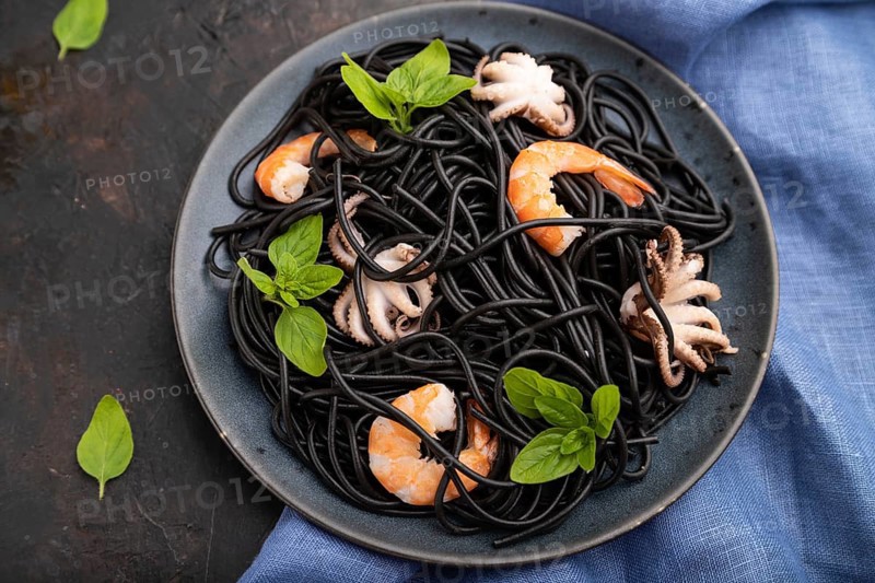 Black cuttlefish ink pasta with shrimps or prawns and small octopuses on black concrete background and blue textile. Top view, close up, selective focus