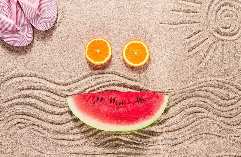 Summer wallpaper, drawing on sand, sea waves and sun, face shape of summer fruits, pink flip-flops, top view, no people