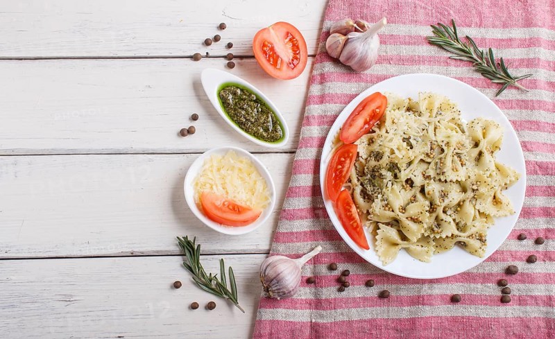 Farfalle pasta with pesto sauce, tomatoes and cheese on a linen tablecloth on white wooden background. top view, copy space
