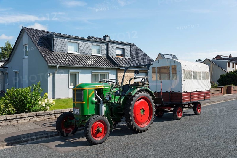 A green tractor with red wheels pulls a white trailer in front of a house under a blue sky, Ilsede, Peine district, Lower Saxony, Germany, Europe