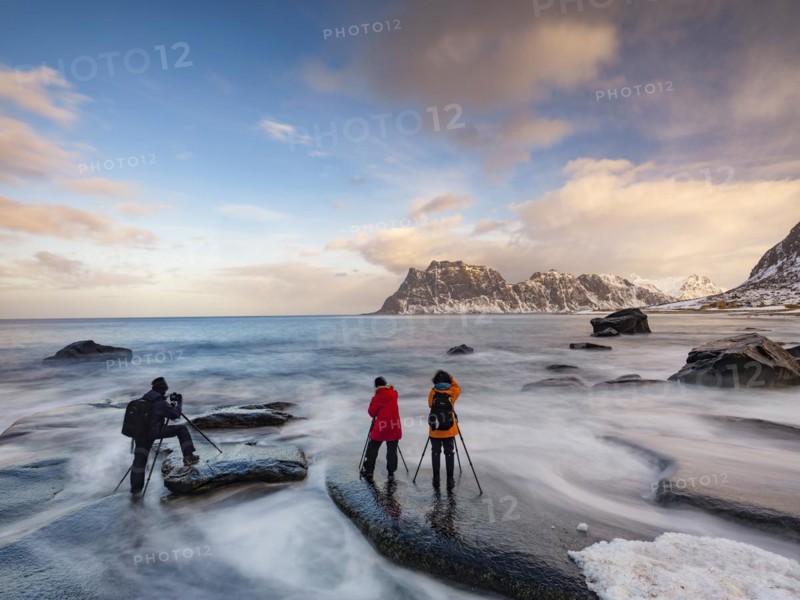 Photographers with tripod on smooth rocks on Utakleiv beach in a dramatic cloudy atmosphere, snow-covered mountains in the background, Vestvagoy Island, Lofoten, Norway, Europe
