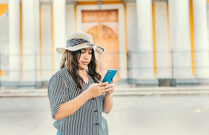 The girl wears a hat, stands in front of a Catholic church with a blurred background, is smiling and sending messages from her cell phone