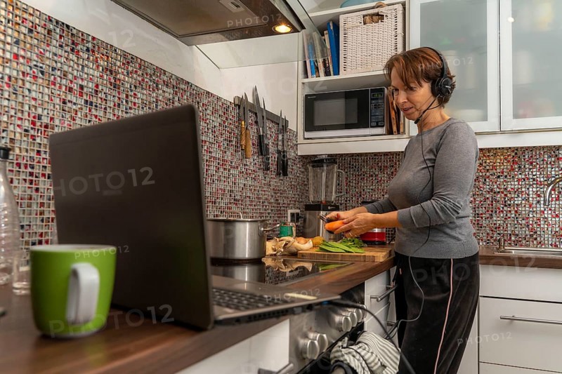 Woman, mid-50s, works from home, with laptop and communicates with colleagues via headset, home office, in the kitchen, while cooking