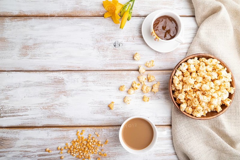 Popcorn with caramel in wooden bowl and a cup of coffee on a white wooden background and linen textile. Top view, flat lay, copy space