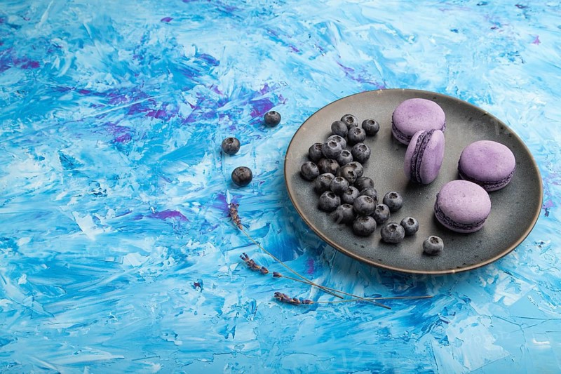 Purple macarons or macaroons cakes with blueberries on ceramic plate on a blue concrete background. Side view, copy space, close up