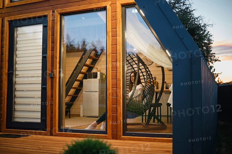 A cozy and stylish tiny house interior captured during twilight. The image showcases a woman lounging comfortably in a unique egg chair, reflecting a serene lifestyle