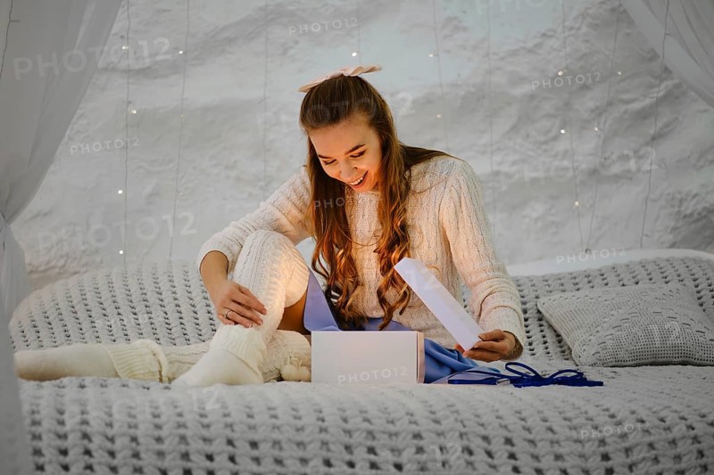 Young woman opens a gift box and admires the light emanating from it. She is sitting on bed and looking inside the glowing box