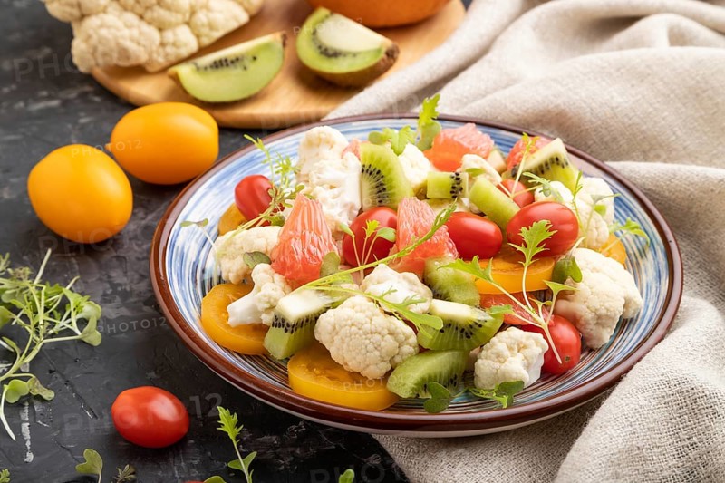 Vegetarian salad of cauliflower cabbage, kiwi, tomatoes, microgreen sprouts on black concrete background and linen textile. Side view, close up