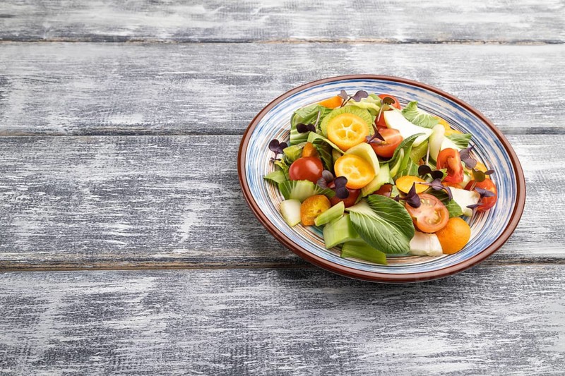 Vegetarian salad of pac choi cabbage, kiwi, tomatoes, kumquat, microgreen sprouts on gray wooden background. Side view, copy space, close up