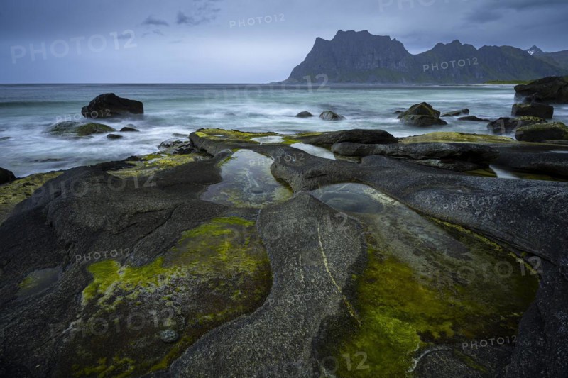Seascape on the beach at Uttakleiv (Utakleiv), in the foreground rocks and the so-called Eye of Uttakleiv, a stone in the water. In the background the mountain Hogskolmen. Cloudy sky. Early summer. Long exposure. Uttakleiv, Vestvagoya, Lofoten, Norway, Europe