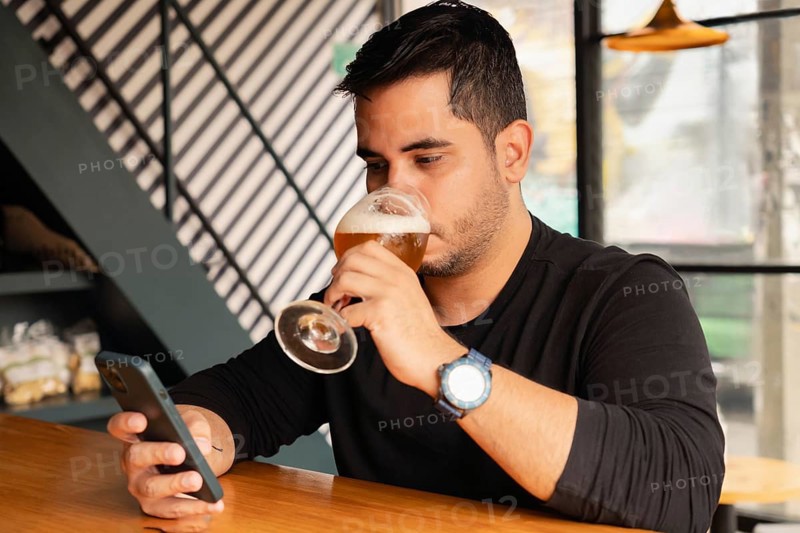 A Latin man in black shirt sits at a wooden bar table, enjoying a glass of beer during the day in a stylish modern bar, entertaining himself by looking at his cell phone