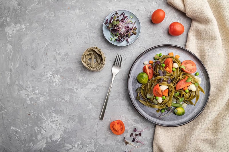 Tagliatelle green spinach pasta with tomato, pea and microgreen sprouts on a gray concrete background and linen textile. Top view, flat lay, copy space