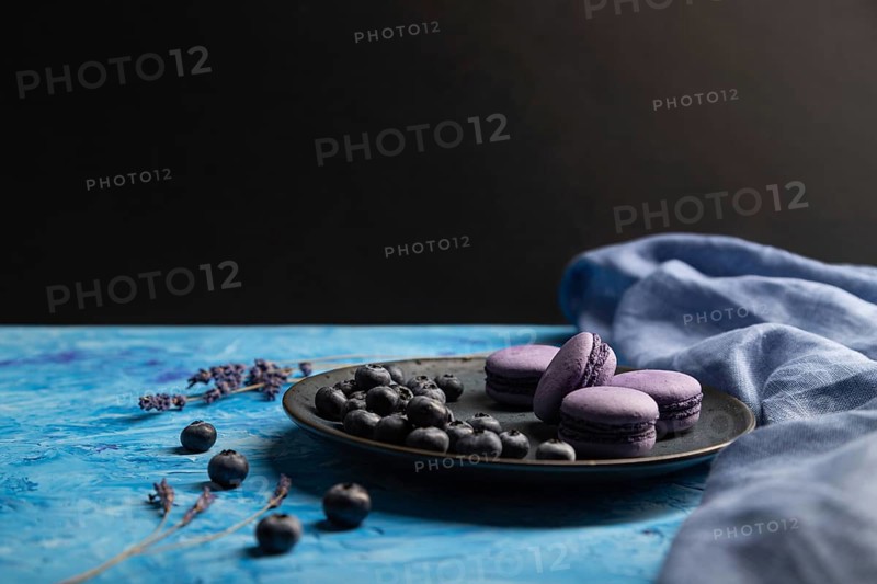 Purple macarons or macaroons cakes with blueberries on ceramic plate on a blue and black background and blue textile. Side view, copy space, selective focus