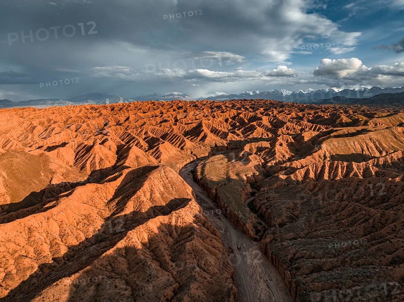 River bed runs through a landscape of eroded hills, badlands at sunset, mountain peaks of the Tian Shan Mountains in the background, aerial view, Canyon of the Forgotten Rivers, Issyk Kul, Kyrgyzstan, Asia