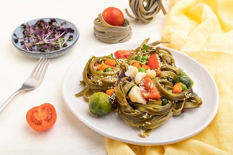 Tagliatelle green spinach pasta with tomato, pea and microgreen sprouts on a white wooden background and yellow textile. Side view, close up