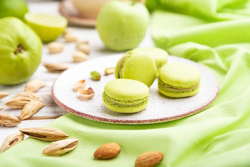 Green macarons or macaroons cakes with cup of coffee on a white wooden background and green linen textile. Side view, close up, selective focus