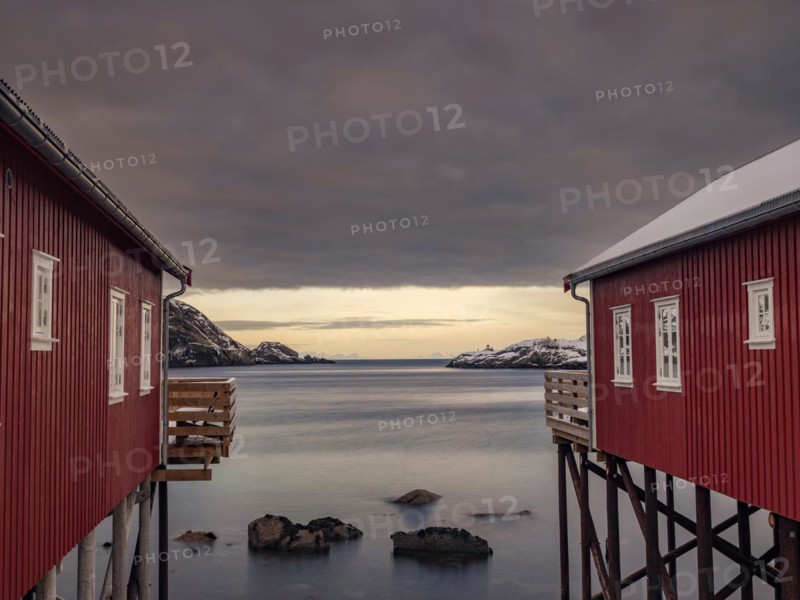 View between two traditional red wooden houses across the fjord to the mountains of the mainland, Nusfjord, Flakstadoya, Lofoten, Norway, Europe