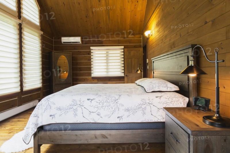 King size bed on wood-frame with wooden headboard, nightstand with antique industrial style lamp, dresser with oval mirror in master bedroom inside a milled Eastern white pine timber and flat log profile home, Quebec, Canada, North America