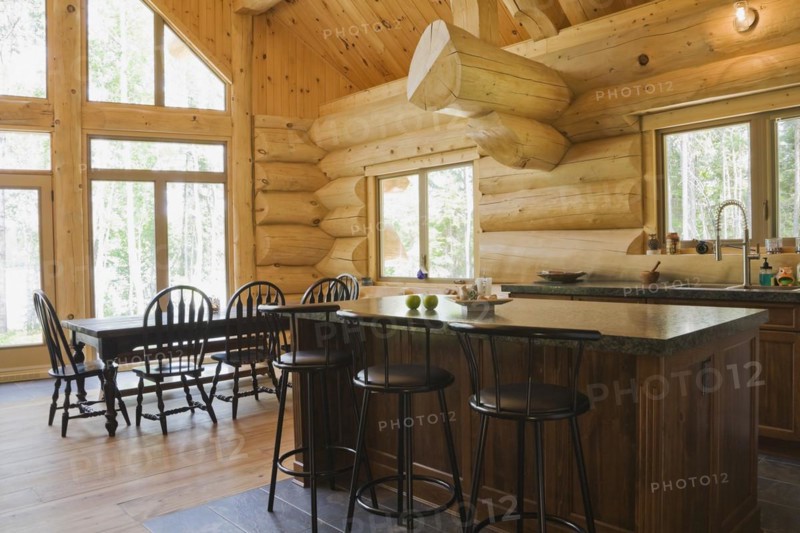 Island with dark grey granite countertop and black stained wooden bar stools plus antique wood table with rounded high back chairs in in open concept kitchen dining room inside handcrafted Eastern white pine Scandinavian log cabin home, Quebec, Canada, North America