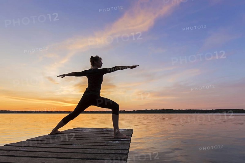 Woman practicing yoga posture Virabhadrasana II, Warrior Pose, lunging standing asana on jetty at lake at sunset