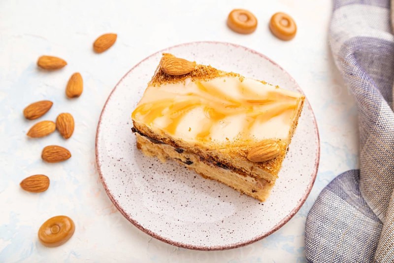 Honey cake with milk cream, caramel, almonds and a cup of coffee on a white concrete background and linen textile. Side view, selective focus, close up