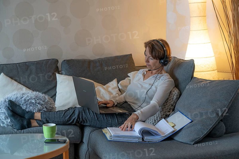 Woman, mid-50s, works from home, with laptop and communicates with colleagues via headset, home office, on the sofa