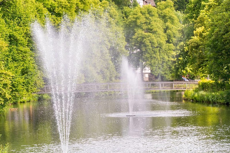 Water fountains bubble in a tranquil lake, surrounded by lush greenery and a bridge path in the background, Stadtpark Kleb, Nagold, Black Forest, Germany, Europe