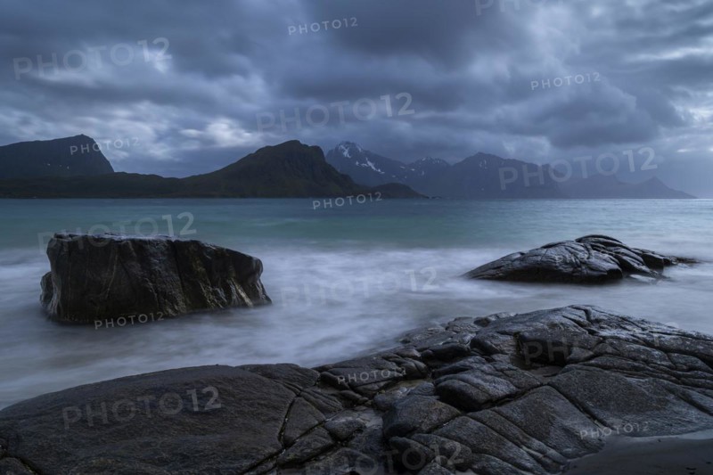 Seascape on the beach at Haukland. View of the mountains of Vestvagoya and Myrland on Flakstadoya. Offersoykammen on the far left. Clouds in the sky. Rocks in the foreground. Early summer. Long exposure. Haukland Beach, Haukland, Vestvagoya, Lofoten, Norway, Europe