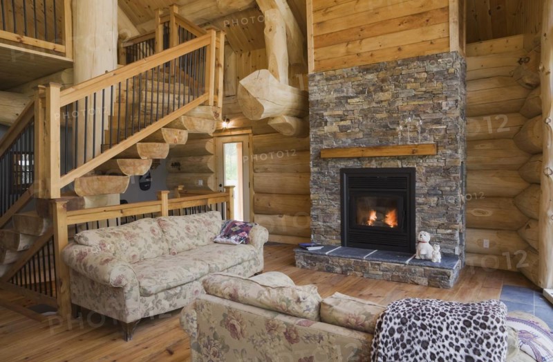 High angle view of twisting staircase with wooden and black wrought iron handrail and brown stained half log steps attached to large supporting central tree trunk post plus lit natural stone fireplace in living room with hardwood floor and flowery motifs upholstered sofas inside handcrafted Eastern white pine Scandinavian log cabin home, Quebec, Canada, North America