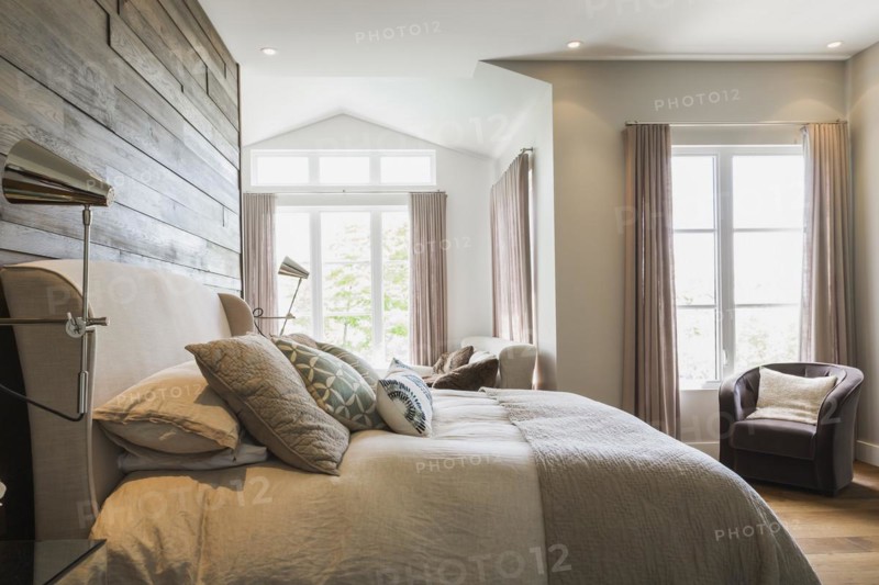 King size bed with grey linen cloth headboard, charcoal velour armchair in master bedroom with engineered hickory wood floorboards inside a luxurious contemporary bungalow style home, Quebec, Canada, North America