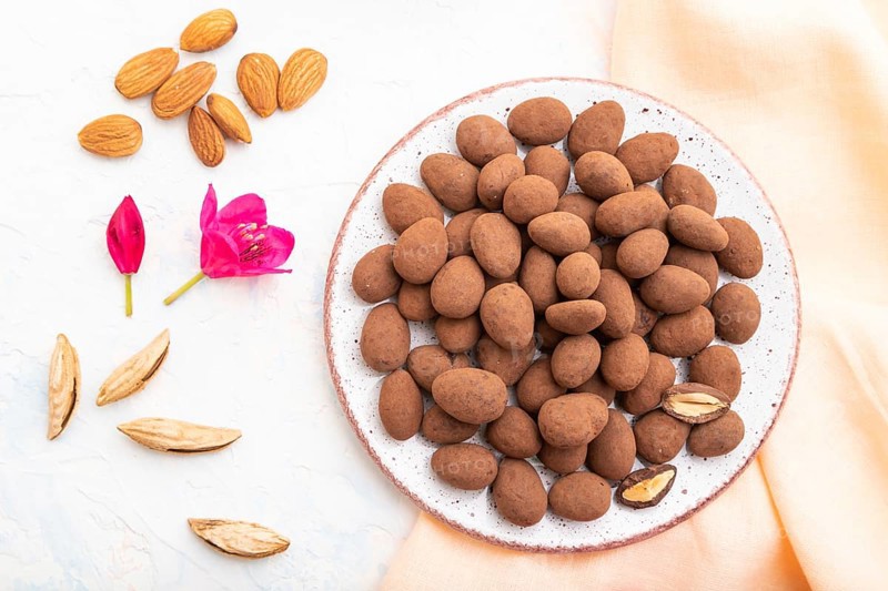 Almond in chocolate dragees on ceramic plate and a cup of coffee on white concrete background and orange linen textile. Top view, close up, flat lay
