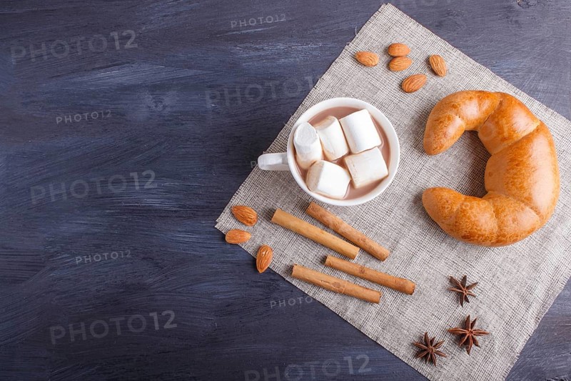 A cup of hot chocolate with marshmallow, croissant and spices on linen napkin and black wooden background. top view, copy space