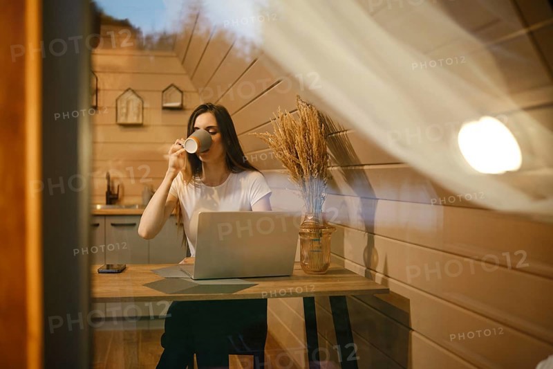 View through the window at a woman sitting at a table and drinking tea in wooden house in the evening