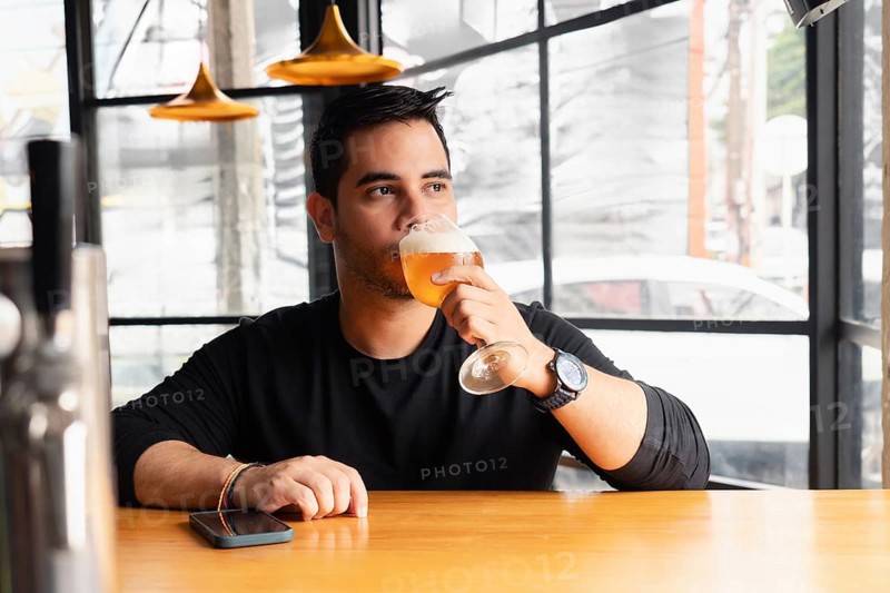 A young Latin man in a black shirt tastes a lager beer at a wooden bar table in a modern bar with stylish decor. The setting features unique lighting and a contemporary design, perfect for relaxation and socializing