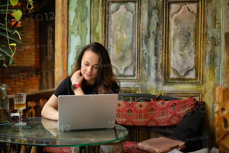 Amidst the rustic charm of a vintage cafe, an adult woman focuses on a laptop, blending modern technology with the cozy ambiance of peeling paint and lush greenery